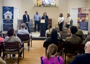 A group of people sitting in a room with banners for Mercy Housing and Shelter Corporation and My Sisters Place.